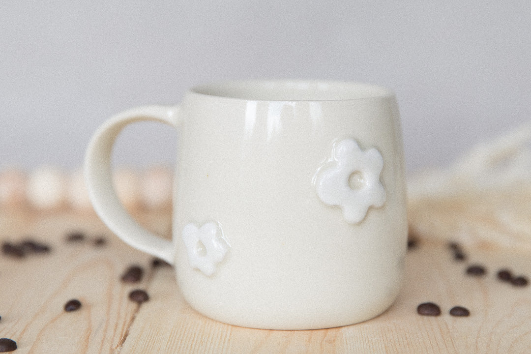 White ceramic mug with white raised flowers on a wooden surface with coffee beans.